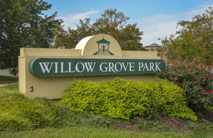 Monument sign for Willow Grove Park in Willow Grove, PA, with green shrubs and rose bushes in front and a brick building behind the trees.